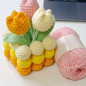 Puede incluir: Un arreglo de tulipanes hechos a mano con ganchillo en tonos de amarillo, naranja, rosa y blanco. El ramo incluye hojas verdes y se combina con una madeja de hilo rosa. El proyecto artesanal es probablemente para la decoración del hogar o un regalo.