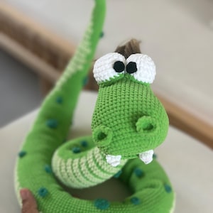 May include: A handmade green and white crochet snake toy with large white eyes and a friendly expression. The snake has a long, curved body with teal accents and a green head. The toy is soft and appears to be stuffed.