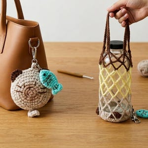 Puede incluir: Un bolso tote de cuero marrón con un llavero de oso de ganchillo y un dije de pez turquesa. Una botella de vidrio transparente se sostiene en un soporte de ganchillo con hilo marrón y amarillo. También se ven un ganchillo y ovillos de lana.