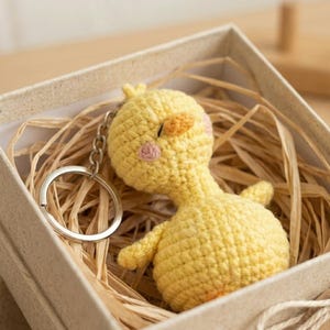 May include: A handmade yellow crochet chick keyring with orange beak and pink cheeks, presented in a beige gift box filled with straw. The keyring has a silver metal ring.