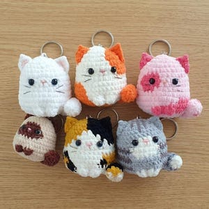 以下が含まれることがあります： さまざまな色の手作りの猫のキーホルダー6個セット。白い猫、オレンジ色の猫、ピンク色の猫、茶色の猫、黒色の猫、灰色の猫が特徴で、黒い目とひげがあります。各猫にはシルバーのキーリングが付いています。