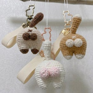 May include: Three handmade crochet cat butt keychains in cream, tan, and white. Each keychain features a cat-shaped charm with a tail and a loop for attaching keys. The keychains are suspended from a wooden branch.