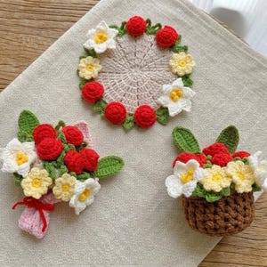 May include: Crocheted floral designs in the image include a bouquet, a basket of flowers, and a coaster. The flowers are red, white, and yellow, with green leaves. The coaster is circular and has a light beige center.