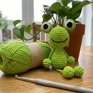 Puede incluir: Una muñeca de rana verde hecha a mano con ojos grandes en blanco y negro, junto a una madeja de hilo verde y un ganchillo. Una planta en maceta está en el fondo. El ganchillo es de 2,6 mm.