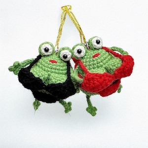 May include: Two handmade crochet frog ornaments. The frogs are green with large white and black eyes, red mouths, and wear black and red swimsuits. They hang from a gold string against a white background.