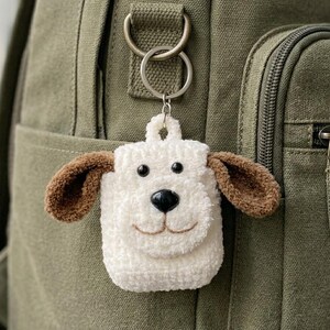 May include: A handmade white and brown dog-shaped keychain. The keychain is made of a soft, textured material with black eyes and a black nose. The dog has brown ears and a smiling mouth. The keychain is attached to a silver keyring.