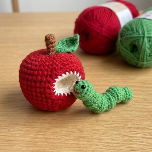 May include: A red crocheted apple with a brown stem and a green leaf, featuring a white bite mark. A small green crocheted worm with black bead eyes and a red stitched mouth emerges from the bite mark. Two balls of yarn are visible in the back.