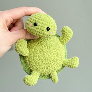 Peut inclure: Une peluche tortue au crochet vert clair, faite à la main, avec un corps, une tête et des membres ronds. La tortue a des yeux noirs et des joues roses. La peluche est tenue dans une main sur un fond neutre.