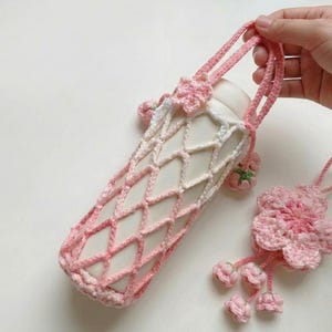 May include: A handmade, pink and white crochet water bottle holder with a matching flower charm. The holder features a diamond pattern and a handle. The charm has a flower design with dangling buds. The items are on a white background.