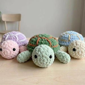 May include: Four handmade crochet turtle plushies in pastel colors. The turtles have textured shells in lavender, green, and blue, with matching limbs and heads. The turtles are displayed on a light wooden surface.