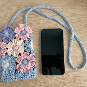 May include: A handmade crochet phone case with a light blue strap. The case is decorated with colorful crochet flowers in shades of pink, blue, purple, and cream, each with a pearl center. A black smartphone is next to the case.