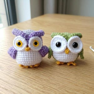 May include: Two handmade crochet owl figurines. One owl is purple and white with yellow eyes, the other is green and white with black eyes. Both have yellow beaks and feet. A crochet hook rests on the wooden surface.