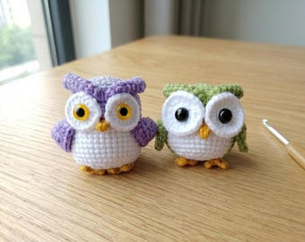 Puede incluir: Dos figuras de búhos de ganchillo hechos a mano. Un búho es morado y blanco con ojos amarillos, el otro es verde y blanco con ojos negros. Ambos tienen picos y patas amarillas. Un ganchillo descansa sobre la superficie de madera.