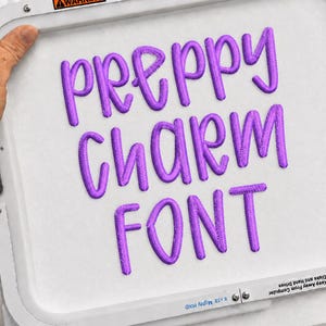 May include: A white embroidery hoop frame containing white fabric with the purple embroidered text PREPPY CHARM FONT. The frame features warning labels about strong magnets and pacemakers, along with text indicating it is an 8x18 Mighty Hoop.