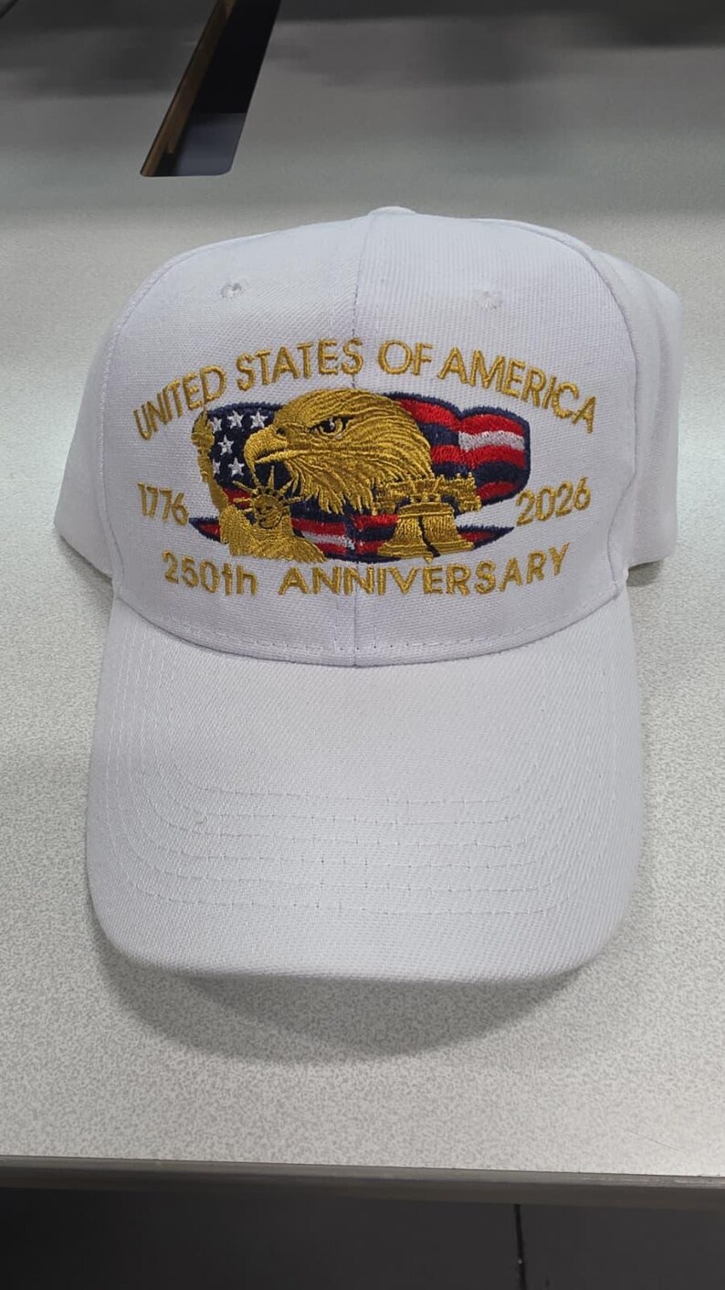 May include: White baseball cap with embroidered text and design. The cap features the words "United States of America" and "250th Anniversary" with the years 1776 and 2026. The design includes an eagle, the American flag, and a bell.