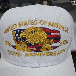 May include: White baseball cap with gold embroidered design. Features the text "UNITED STATES OF AMERICA" and "250th ANNIVERSARY 2026". Includes an eagle, American flag, and the Statue of Liberty.