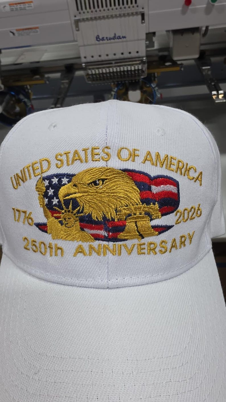 May include: White baseball cap with gold embroidered design. Features the text "UNITED STATES OF AMERICA" and "250th ANNIVERSARY 2026". Includes an eagle, American flag, and the Statue of Liberty.