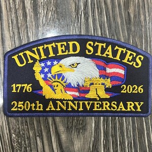 May include: A navy blue patch with gold and white text and imagery. The patch reads "UNITED STATES" above an eagle and American flag design. Below the eagle are the numbers "1776" and "2026" with "250th ANNIVERSARY" below that. The patch also features the Statue of Liberty and the Liberty Bell.