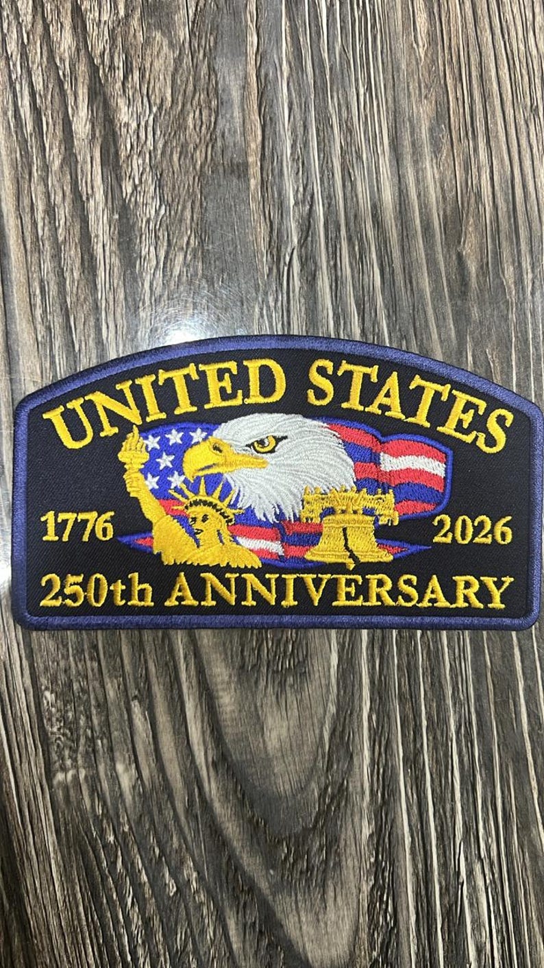 May include: A navy blue patch with gold and white text and imagery. The patch reads "UNITED STATES" above an eagle and American flag design. Below the eagle are the numbers "1776" and "2026" with "250th ANNIVERSARY" below that. The patch also features the Statue of Liberty and the Liberty Bell.
