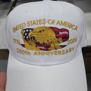 May include: White baseball cap embroidered with "UNITED STATES OF AMERICA" and "250th ANNIVERSARY" in gold. Features an eagle, American flag, and the years 1776 and 2026. A patriotic accessory.