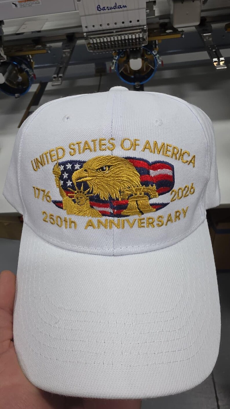 May include: White baseball cap embroidered with "UNITED STATES OF AMERICA" and "250th ANNIVERSARY" in gold. Features an eagle, American flag, and the years 1776 and 2026. A patriotic accessory.