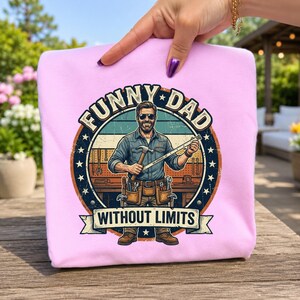 May include: A light pink sweatshirt featuring a circular graphic of a smiling person wearing sunglasses, a denim shirt, and a tool belt. The text reads "FUNNY DAD" and "WITHOUT LIMITS" with a workshop background, a hammer, and a ruler.