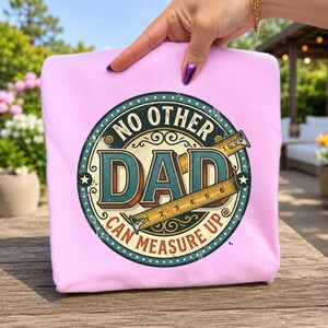 May include: A light pink sweatshirt featuring a circular graphic with a vintage-style design. The text reads NO OTHER DAD CAN MEASURE UP, centred around the word DAD, with a yellow measuring tape graphic showing inches across the centre.