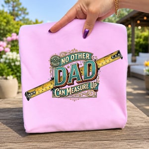 May include: A light pink t-shirt featuring a graphic design of a yellow measuring tape crossing behind the text "NO OTHER DAD CAN MEASURE UP" in teal and brown. A small circular emblem with a hammer icon and the text "WORLD'S BEST FATHER'S DAY" is visible.