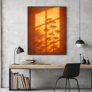 Puede incluir: Una obra de arte enmarcada con una pared de tonos cálidos anaranjados que muestra la silueta de una planta y la luz de una ventana. La obra de arte está sobre un escritorio de madera con una silla negra, una lámpara y libros, creando una escena moderna de oficina o decoración del hogar.
