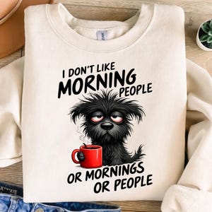 Op de afbeelding: Crèmekleurige sweatshirt met de tekst "I DON'T LIKE MORNING PEOPLE OR MORNINGS OR PEOPLE". De afbeelding toont een slaperige zwarte hond met een rode koffiemok. De sweatshirt heeft een ronde hals.