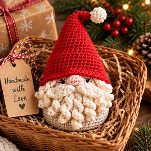May include: A crocheted Santa Claus gnome with a red pointed hat, a white pom-pom, and a curly cream-colored beard. A brown paper tag attached with red and white twine features the text "Handmade with Love" and a small heart icon.