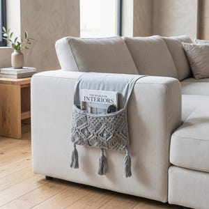 Pode incluir: Um organizador de braço de sofá em tecido cinza claro com um bolso de macramé com padrão de diamante e três borlas pendentes. O bolso contém uma revista intitulada The World of Interiors e óculos pretos sobre um sofá bege.