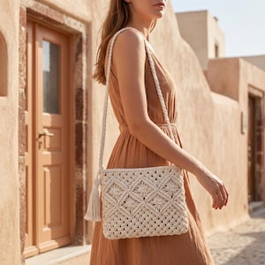 May include: A cream-colored macrame crossbody bag featuring a geometric diamond pattern and a braided shoulder strap. The bag is finished with a long, matching tassel attached to the side, showcasing intricate knotted cord construction throughout.