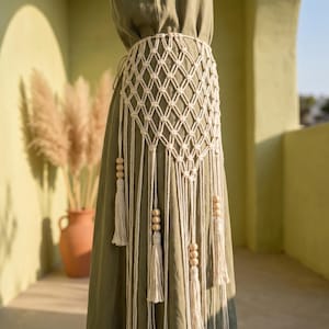 May include: An off-white macrame belt featuring a diamond-shaped knotted lattice pattern. The belt includes long hanging cords finished with wooden beads and tassels, worn over a solid olive green garment that reaches the floor.