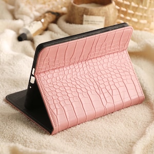 May include: A tablet case featuring a light pink exterior with a textured crocodile skin pattern. The case is designed to fold into a stand, revealing a black interior frame that holds the device securely in place while maintaining a slim profile.