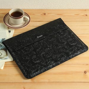 Op de afbeelding: Een zwarte leren laptoptas met een reli&euml;f bloemmotief, het woord "Evans" is in de flap ge&euml;mbosseerd. Een kopje thee op een schotel en een krant liggen op een houten oppervlak.