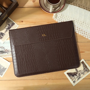 May include: A brown leather laptop sleeve with a crocodile texture, featuring the gold initials "GL". The sleeve is on a wooden surface, alongside vintage postcards, a watch box, and a cup of coffee.