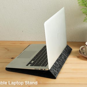 Op de afbeelding: Een zilveren laptop staat op een zwarte, patroon laptopstandaard. De standaard kantelt de laptop voor een betere weergave. De afbeelding staat op een houten oppervlak, met een kopje koffie en een kleine plant op de achtergrond. De tekst "Portable Laptop Stand" staat onderaan.