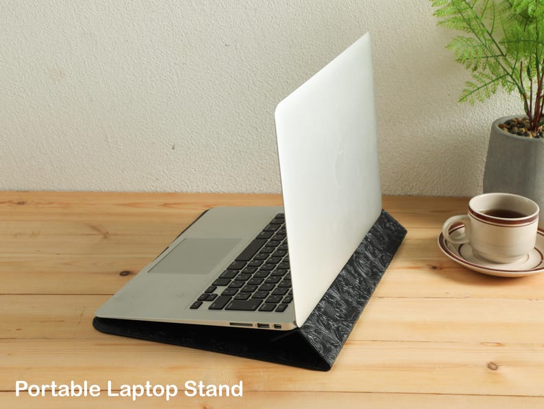 Op de afbeelding: Een zilveren laptop staat op een zwarte, patroon laptopstandaard. De standaard kantelt de laptop voor een betere weergave. De afbeelding staat op een houten oppervlak, met een kopje koffie en een kleine plant op de achtergrond. De tekst "Portable Laptop Stand" staat onderaan.
