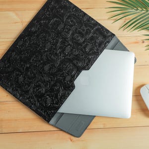 Op de afbeelding: Zwarte laptoptas met een reli&euml;f bloemenpatroon. Een zilveren laptop zit gedeeltelijk in de hoes. Een witte draadloze muis ligt naast de laptop en de hoes. De items liggen op een houten oppervlak.