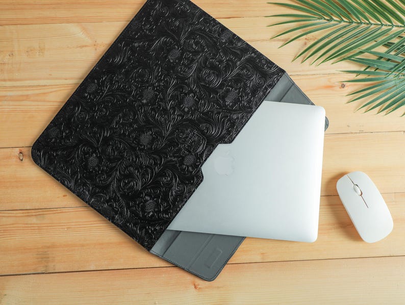 Op de afbeelding: Zwarte laptoptas met een reli&euml;f bloemenpatroon. Een zilveren laptop zit gedeeltelijk in de hoes. Een witte draadloze muis ligt naast de laptop en de hoes. De items liggen op een houten oppervlak.