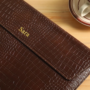 Op de afbeelding: Een bruine leren clutch met een krokodillenstructuur. De naam "Sara" is in gouden letters op de voorkant ge&euml;mbosseerd. De clutch is gesloten en ligt op een houten oppervlak.