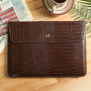 May include: A brown leather laptop sleeve with a crocodile texture. The sleeve has a flap closure and the initials "AK" in gold. A newspaper with the headline "HAPPY INDEPENDENCE" is visible in the background.