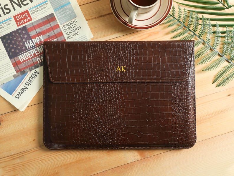 Op de afbeelding: Een bruine leren laptoptas met een krokodillenstructuur. De tas heeft een klepsluiting en de initialen "AK" in goud. Op de achtergrond is een krant met de kop "HAPPY INDEPENDENCE" te zien.