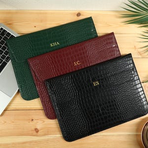 May include: Three personalized laptop sleeves in green, burgundy, and black, with a textured crocodile pattern. Each sleeve has gold-colored initials. A laptop and a cup of coffee are also visible on a wooden surface.