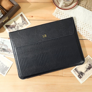 May include: A black leather laptop sleeve with a textured finish and the initials "LB" in gold. The sleeve is on a wooden surface, with vintage postcards and a watch display case nearby.
