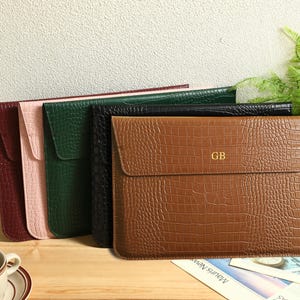 May include: A collection of leather laptop sleeves in various colors, including burgundy, pink, green, black, and brown. The brown sleeve features a crocodile texture and gold initials. A cup of tea sits in the foreground.