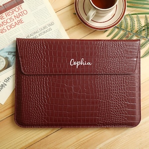 May include: A burgundy laptop sleeve with a crocodile skin texture. The word "Cophia" is embroidered on the front. The sleeve is rectangular with a flap closure. A newspaper and a cup of tea are in the background.