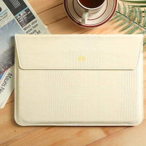 Op de afbeelding: Cr&egrave;mekleurige laptoptas met een krokodillenleertextuur. De klepsluiting heeft de initialen "FB" in goud. Een krant en een kopje koffie staan op de achtergrond.