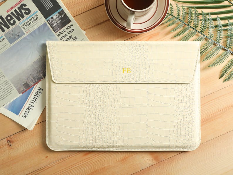 Op de afbeelding: Cr&egrave;mekleurige laptoptas met een krokodillenleertextuur. De klepsluiting heeft de initialen "FB" in goud. Een krant en een kopje koffie staan op de achtergrond.
